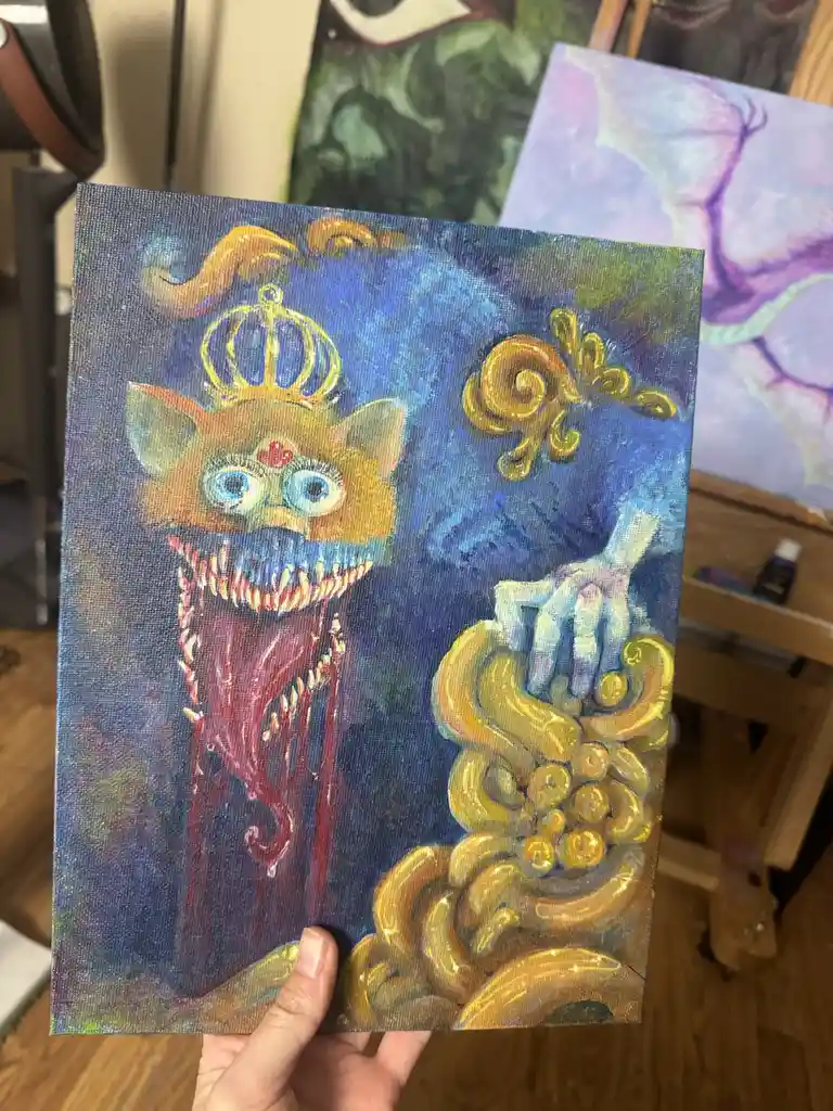 A hand holds up a small canvas painting depicting a cartoonish, orange cat-like monster with large blue eyes, a red heart on…