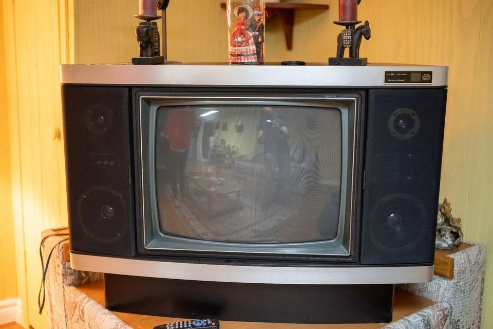 An old, boxy CRT television with integrated speakers sits on a wooden stand, its screen dark and reflective, showing faint…