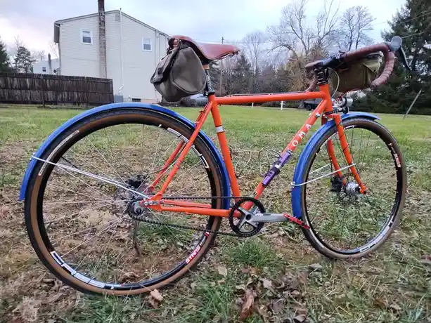 A bright orange bicycle with blue fenders and brown tires is parked on grass, featuring saddlebags and handlebar bags. The…