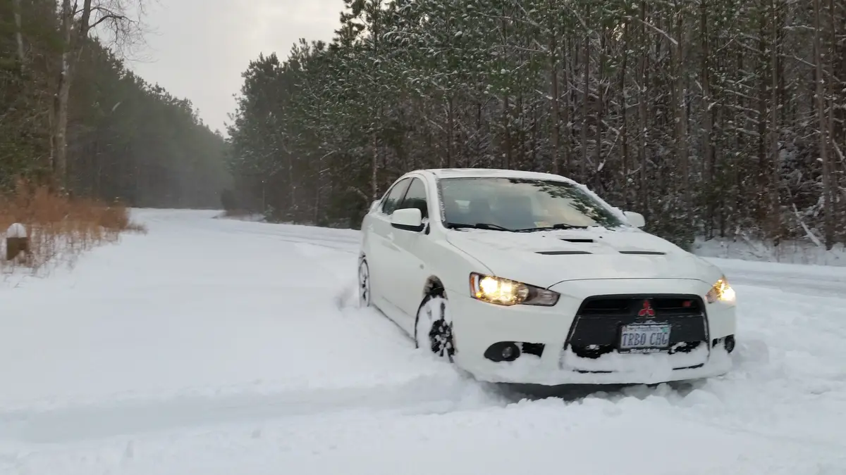 A white Mitsubishi Lancer Evolution is stuck in deep snow on a forest road, its headlights on and snow covering the hood and…