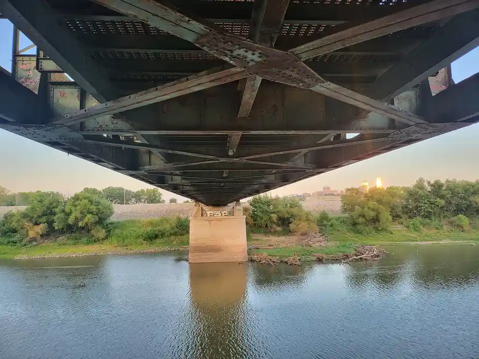 A low-angle photo looking up at the rusty, weathered underside of a large steel bridge spanning a river, with a concrete…