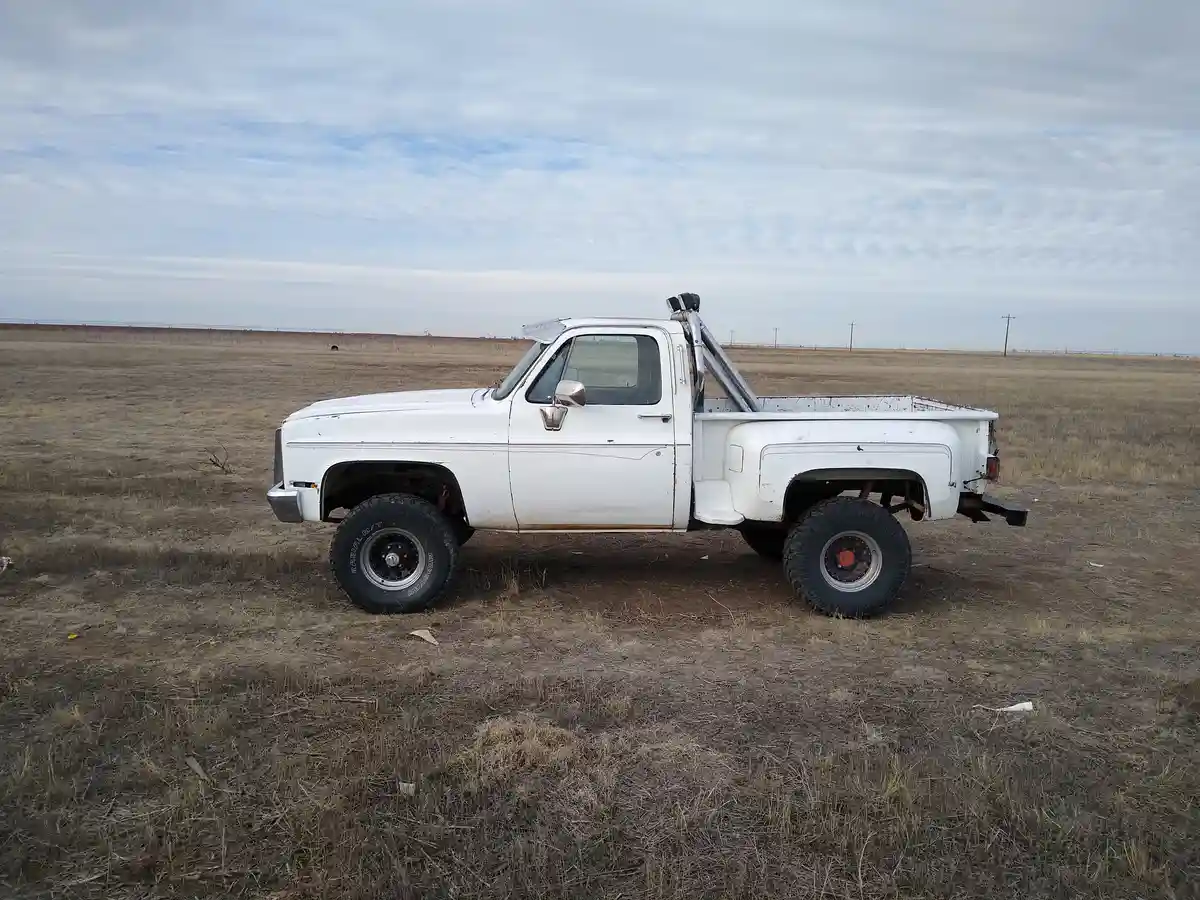 A white, lifted, rusty pickup truck with large off-road tires and a winch sits alone in a vast, dry grassy field under a…