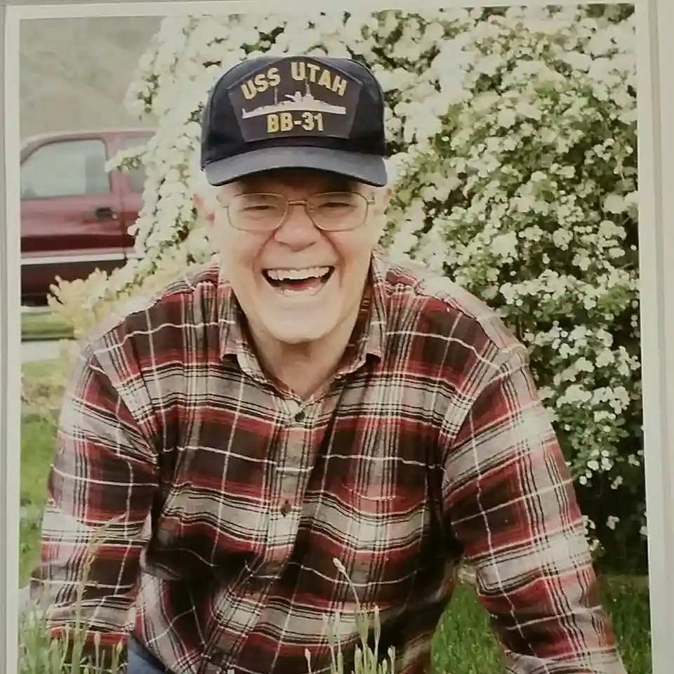 An older man with glasses and a red plaid shirt is laughing joyfully while wearing a navy blue baseball cap that reads 'USS…