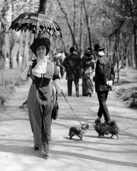 A vintage black-and-white photo shows a woman in early 20th-century attire, holding an ornate parasol and walking two small…