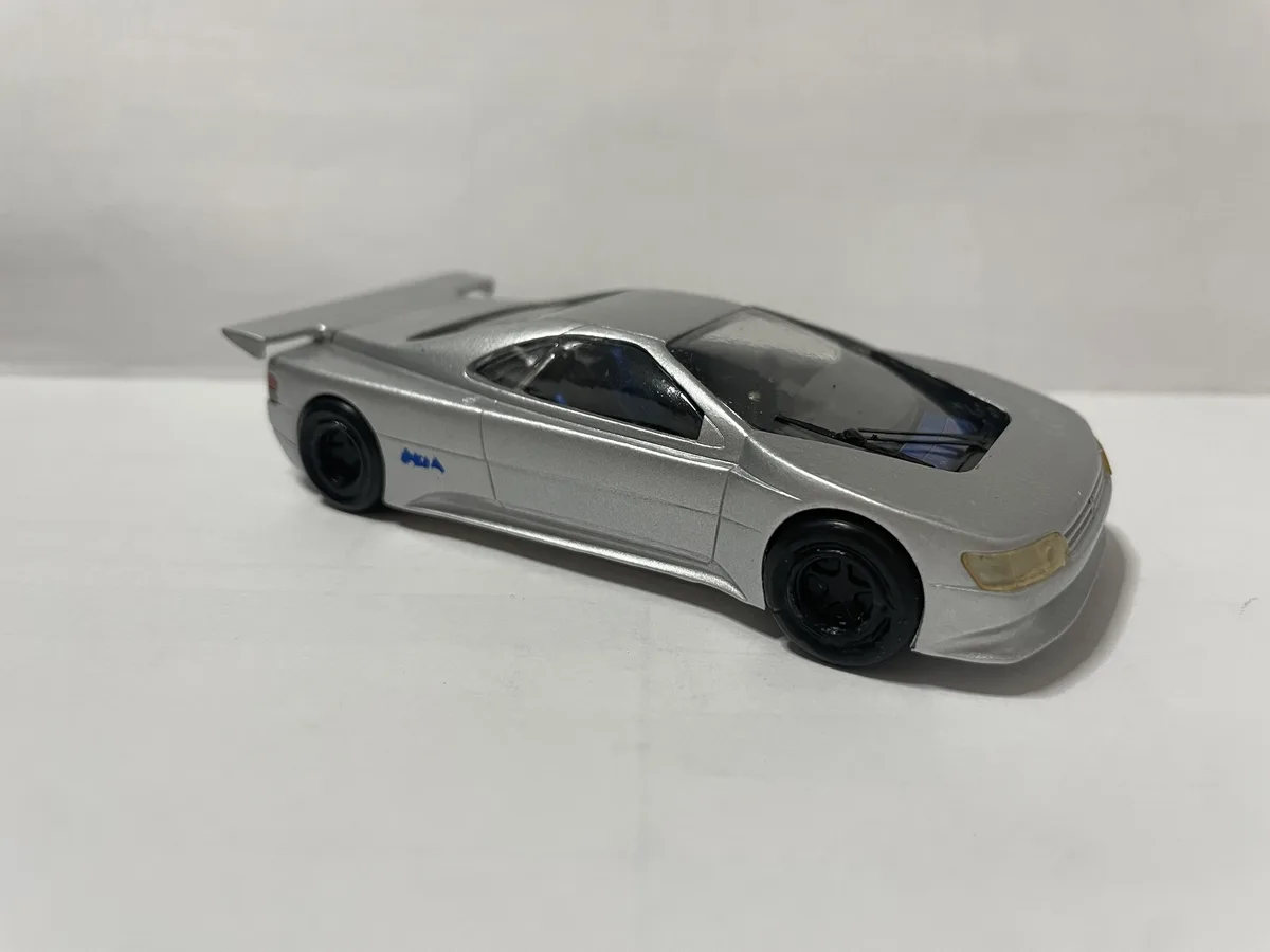 A silver toy car with a large rear spoiler and black wheels is photographed against a plain white background. The car has a…