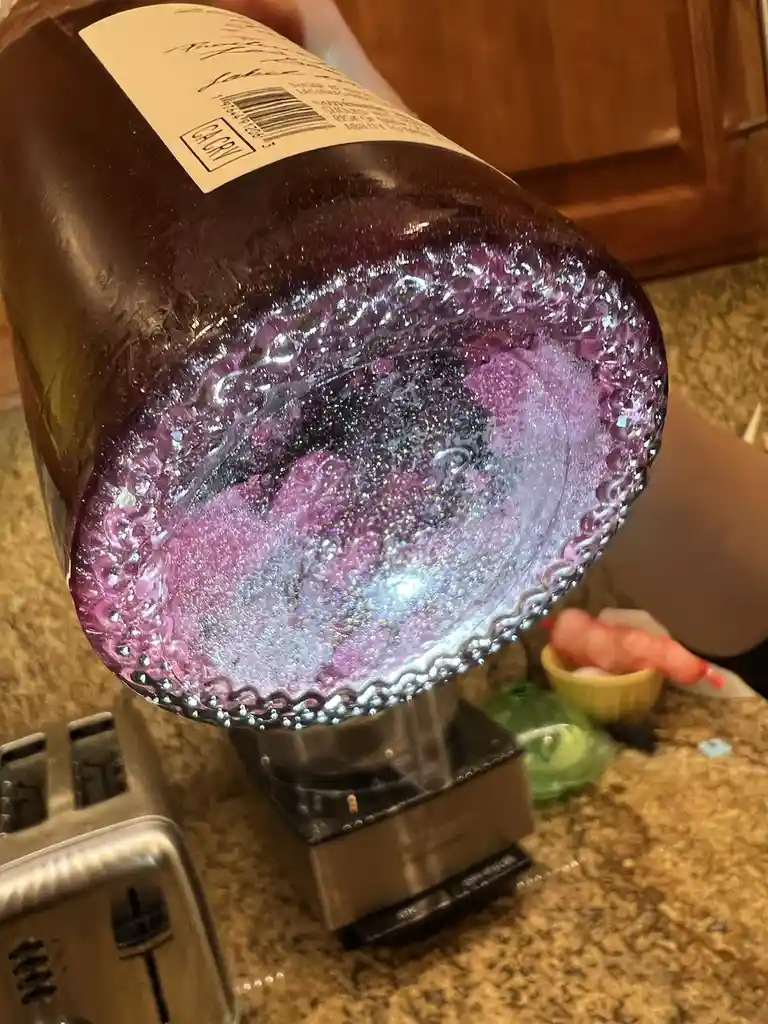 A close-up photo shows the bottom of a dark purple glass bottle, tilted over a kitchen counter, revealing a glittery…