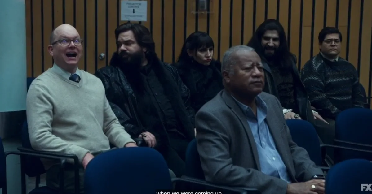 A group of people sit in an audience, with the man on the far left wearing glasses and a light sweater, mouth wide open in…