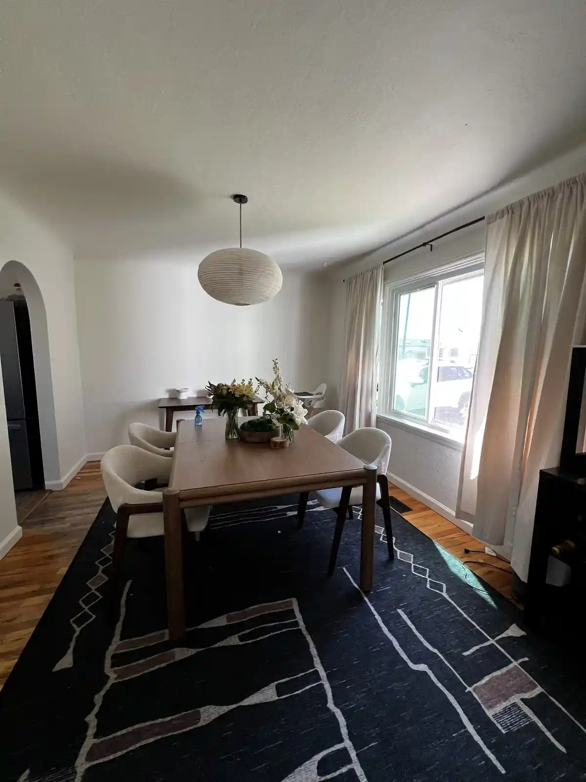 A clean, modern dining room with a wooden table, four white chairs, and a large patterned rug. A white paper lantern hangs…