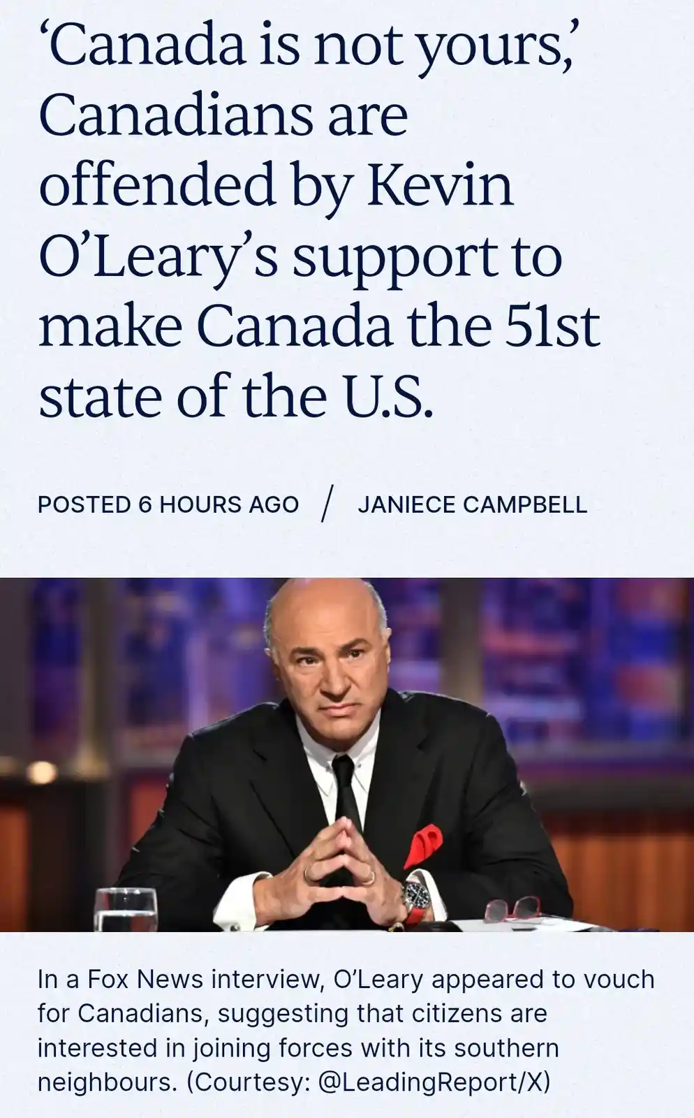 The image shows a man in a black suit with a red pocket square, sitting at a desk with clasped hands, looking serious. The…