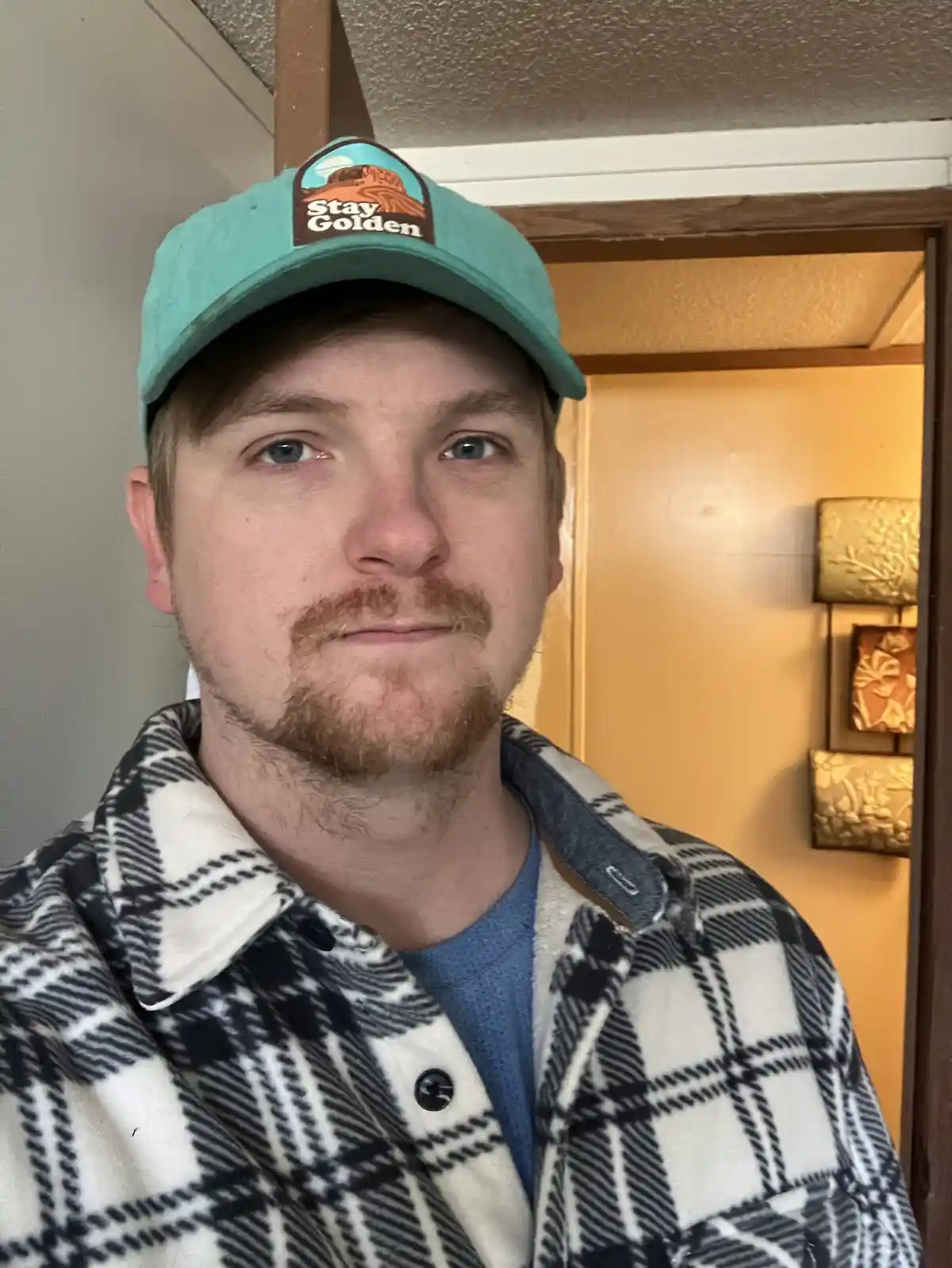 A man with light brown hair and a goatee wears a teal baseball cap with a 'Stay Golden' logo and a black-and-white plaid…
