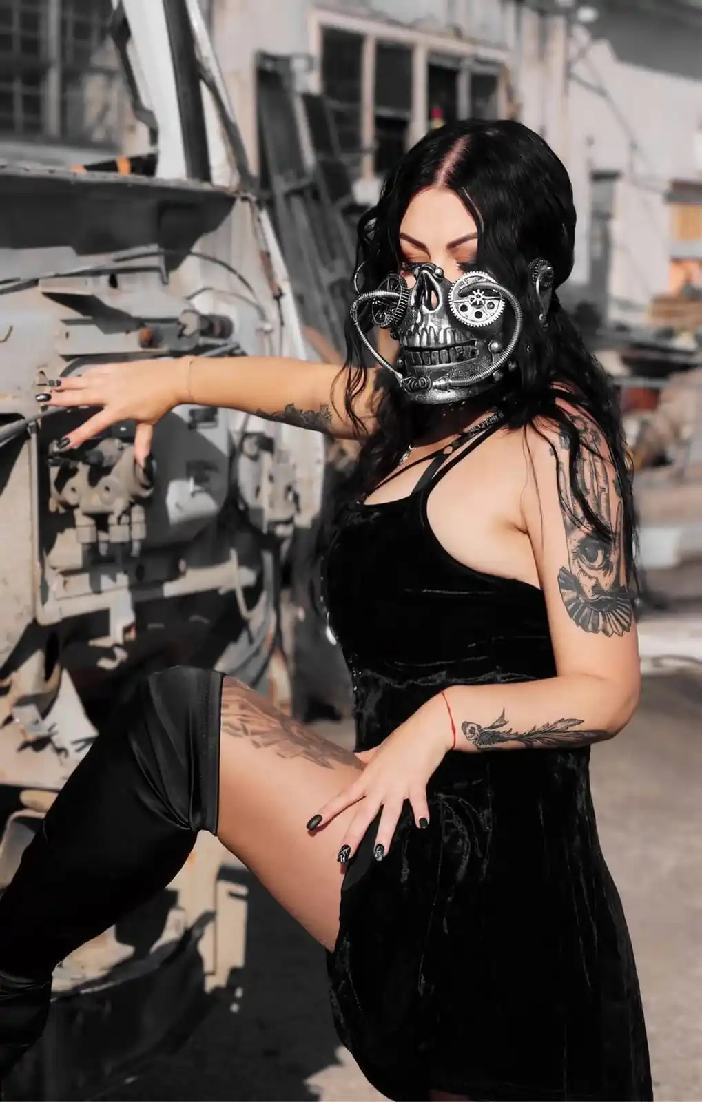 A woman with long black hair and tattoos poses in a black velvet dress and thigh-high boots next to a large, rusted…