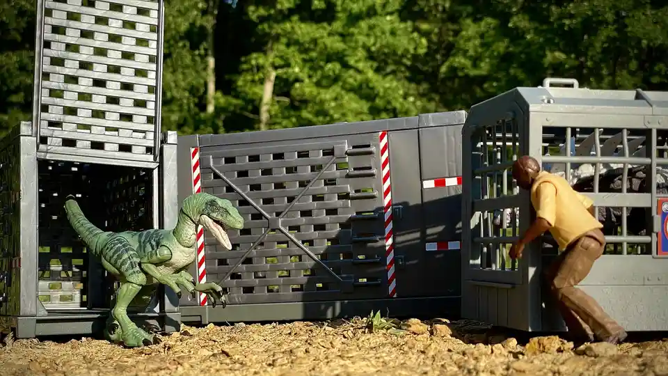 A green toy dinosaur stands outside an open gray plastic cage, facing a toy human figure in a yellow shirt who is leaning…
