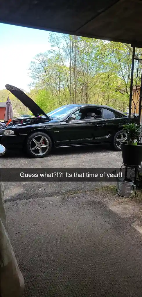A black Ford Mustang with its hood open is parked under a carport, showing its engine bay with tools and wires. The car has…