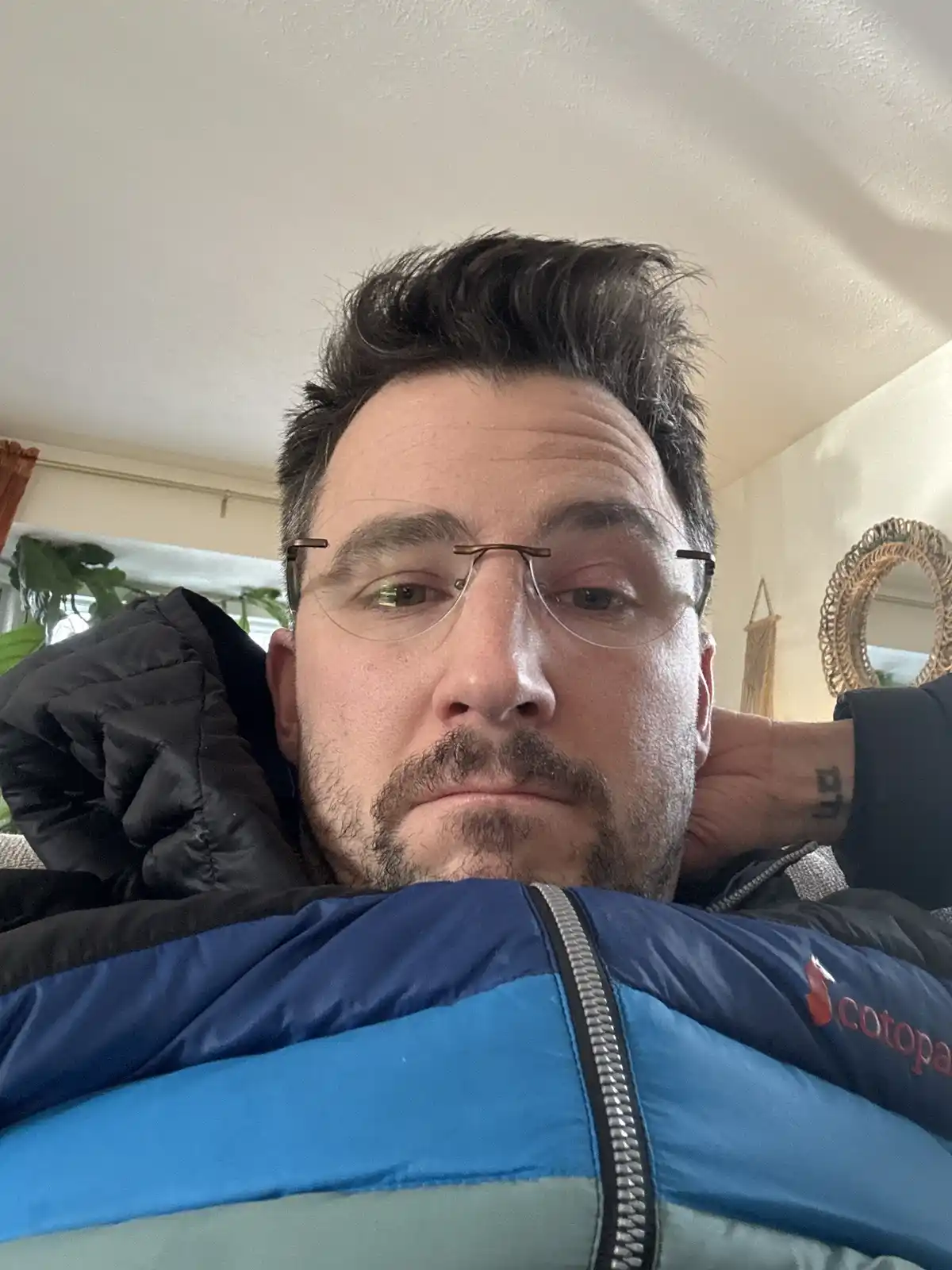 A man with glasses and a mustache is lying down wearing a thick blue and black puffer jacket, looking directly at the camera…