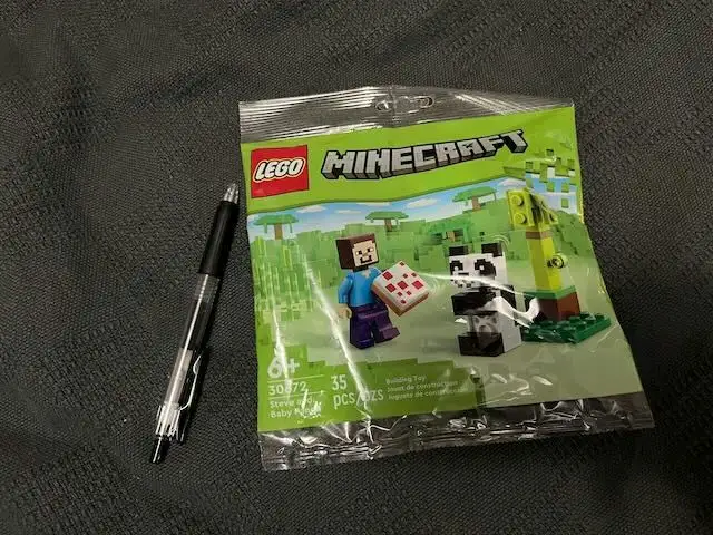 A sealed LEGO Minecraft set (30172) featuring Steve and a baby panda minifigure, shown next to a black pen on a dark gray…