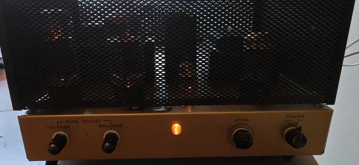 A close-up of a vintage-style tube amplifier with a black perforated metal grille and a tan control panel. The panel…