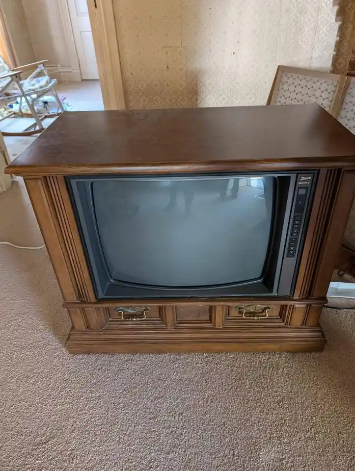 A vintage wooden television set with a curved CRT screen sits on a carpeted floor in a room with patterned wallpaper and a…