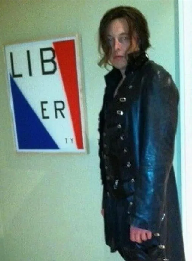 A person in a dark, shiny, buttoned coat stands beside a framed sign with the word 'LIBERTY' split across red and blue…