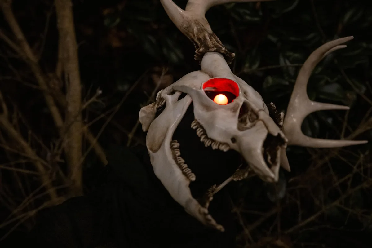 A deer skull with large antlers is shown hanging in a dark, wooded area at night. One eye socket glows with a bright red…