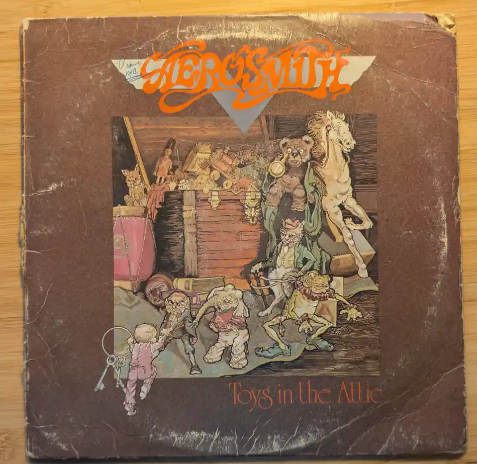 A worn, brown vinyl record album cover for Aerosmith's 'Toys in the Attic' shows a chaotic attic scene with animated toys…