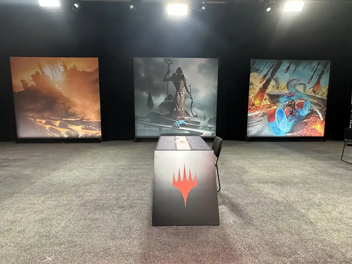 An empty Magic: The Gathering tournament table with the red mana symbol on its front, positioned in front of three large…