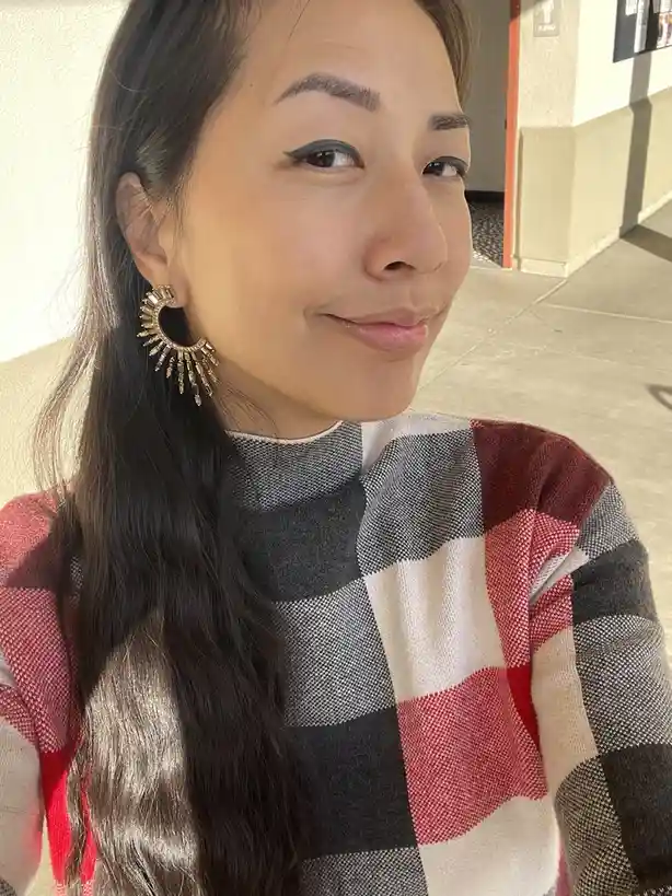 A woman with long dark hair and a red, black, and white plaid sweater smiles while looking slightly away from the camera…