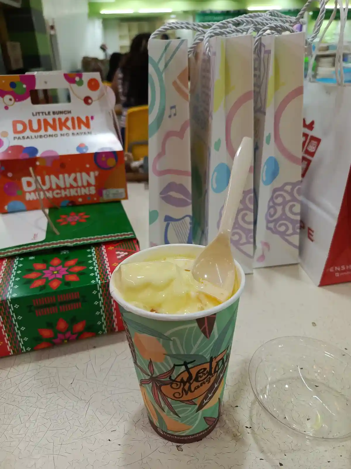 A cup of yellow mango dessert with a spoon sits on a table, surrounded by colorful gift bags and a Dunkin' Munchkins box…