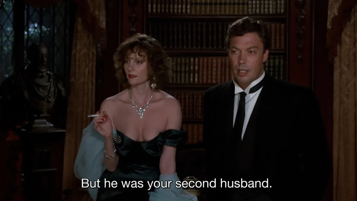 A still from a movie shows a woman in a black dress holding a cigarette and a man in a tuxedo standing beside her in a…