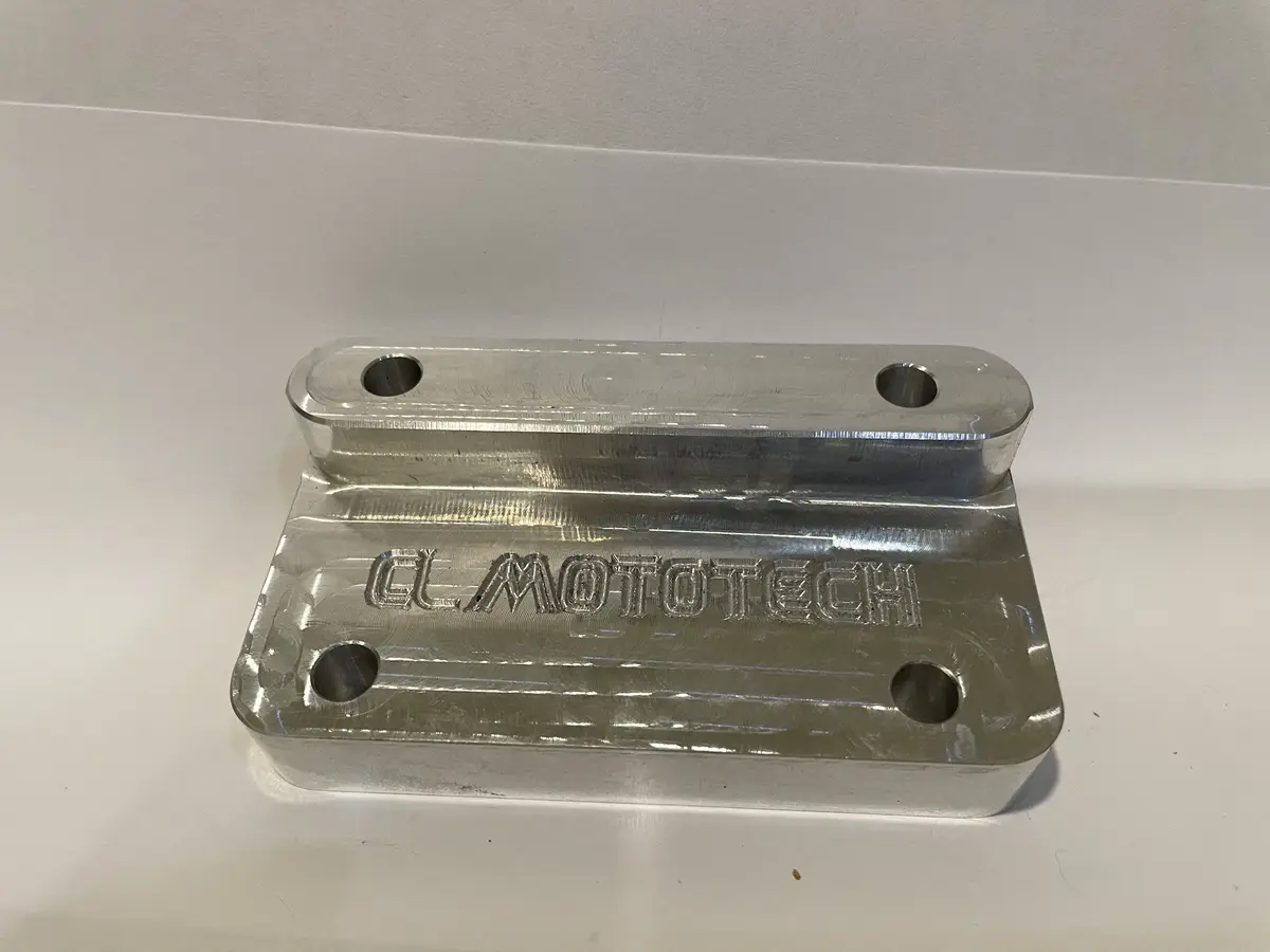 A shiny, silver-colored metal bracket with four bolt holes and the engraved text 'CL MOTOTECH' on its surface, resting on a…