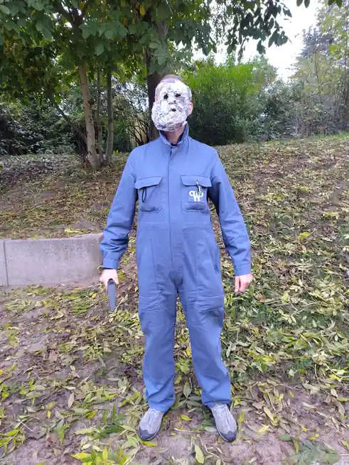 A person stands outdoors in a blue jumpsuit, wearing a makeshift foil mask covering their face and holding a knife in one…