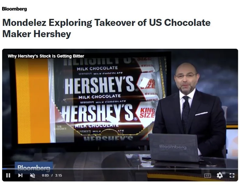 A Bloomberg news anchor sits at a desk in front of a screen displaying a Hershey's chocolate bar package. The overlaid text…
