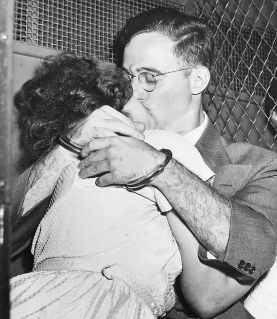 A black-and-white historical photo shows a man in a suit and glasses kissing a woman in a light dress while both are…