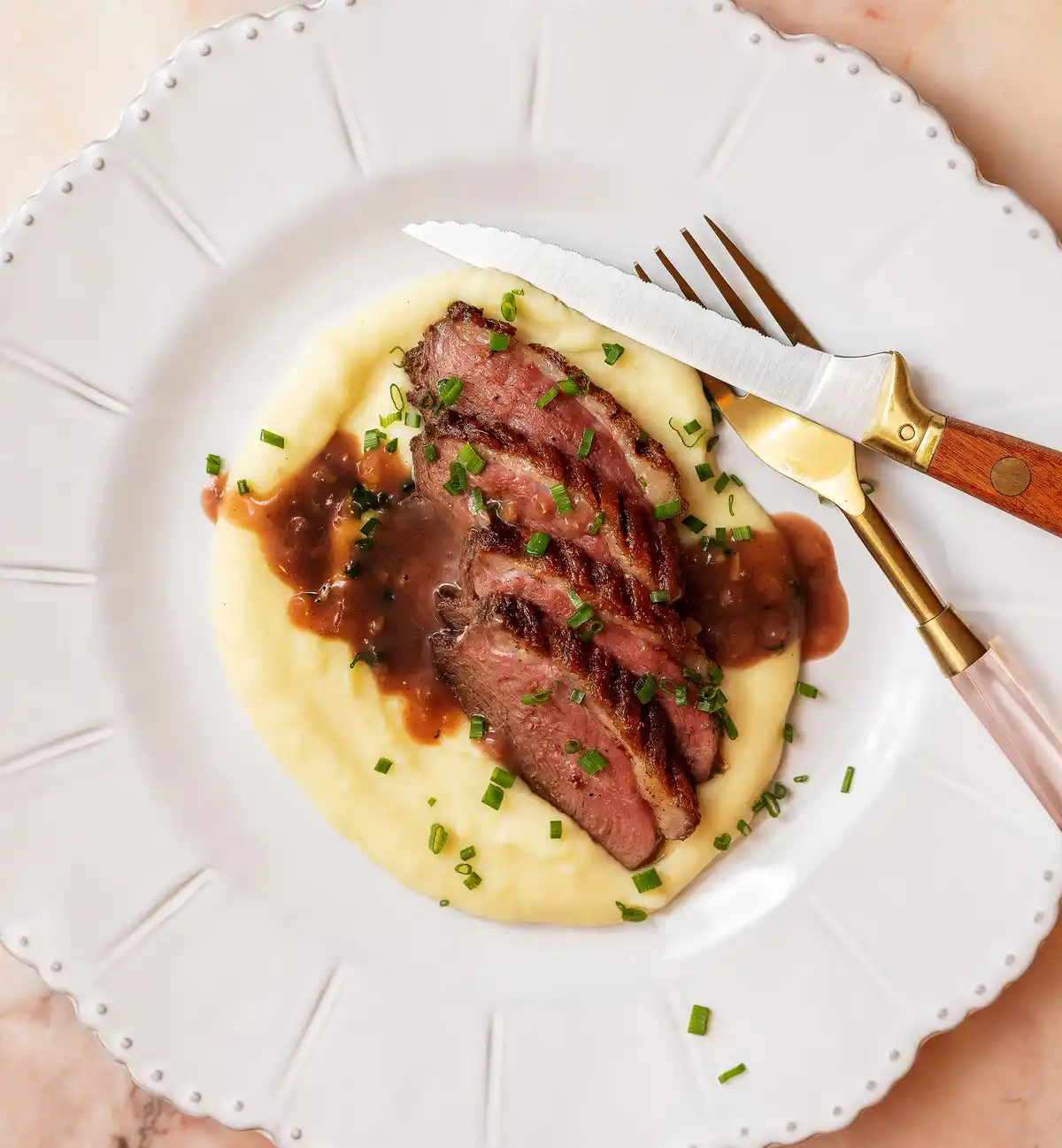 A top-down photo of a plated meal featuring sliced medium-rare meat atop creamy mashed potatoes, garnished with chopped…