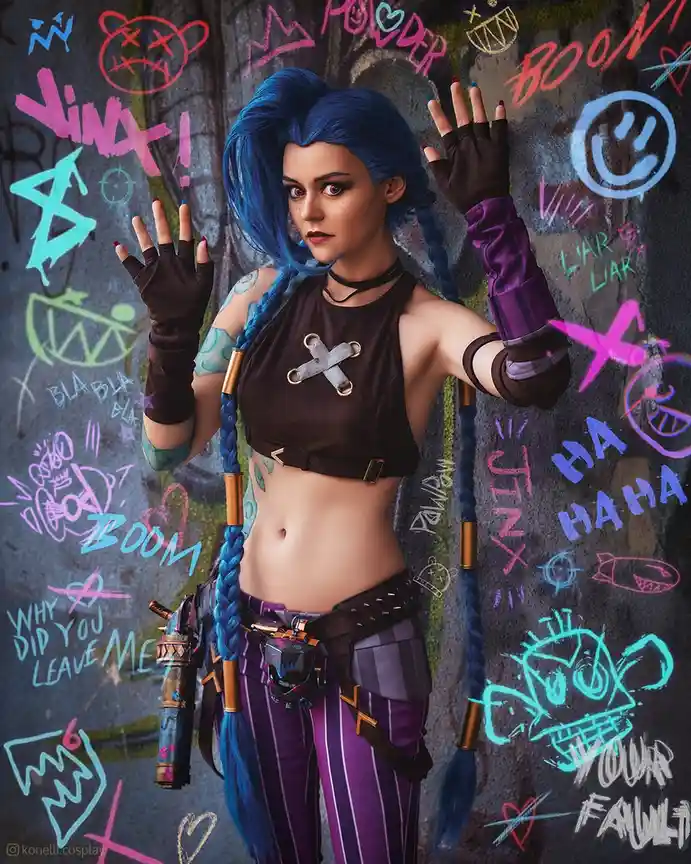 A cosplayer dressed as the character Lina from League of Legends stands with hands raised against a graffiti-covered wall…