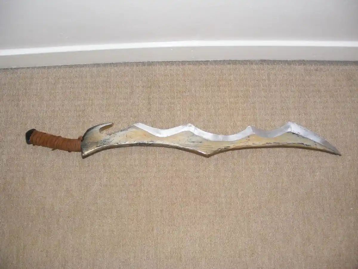 A homemade, wavy-bladed sword with a brown wrapped handle lies horizontally on a beige carpet, with a white baseboard…