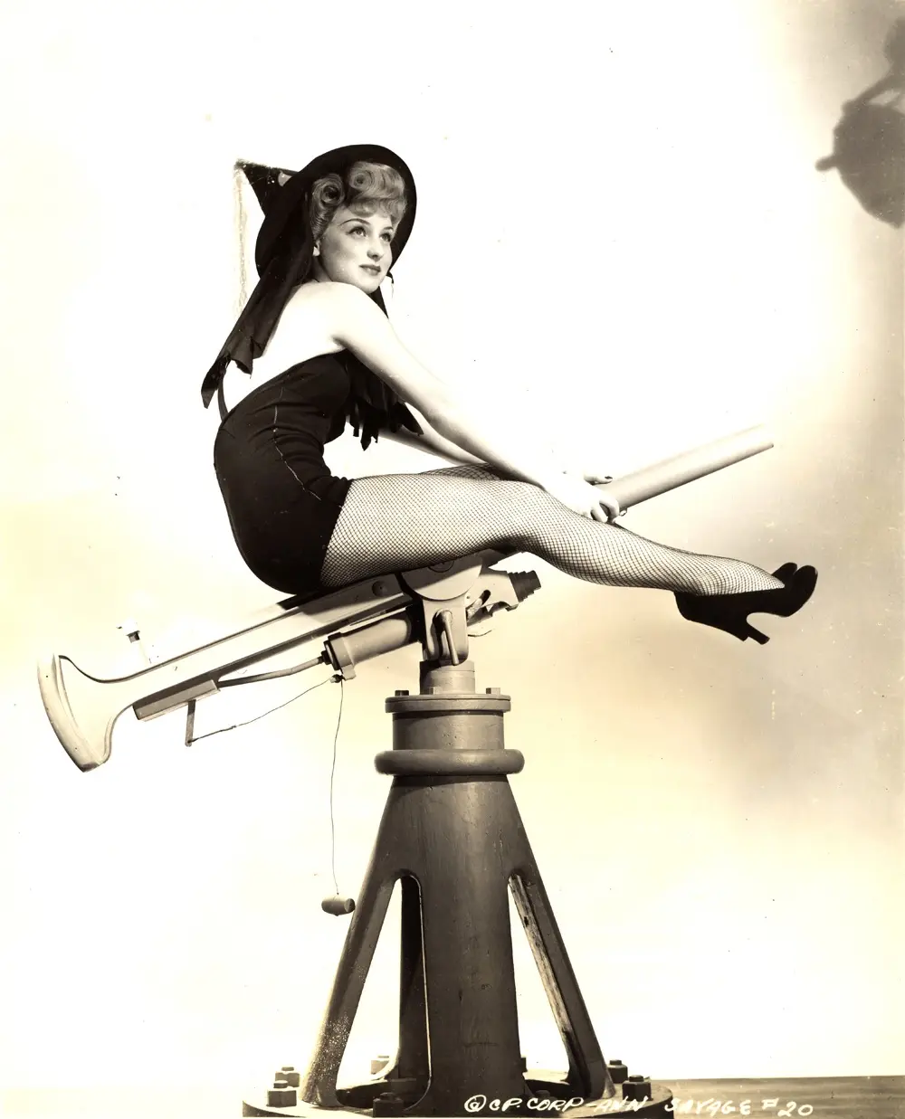 A vintage black-and-white photo shows a woman in a witch costume, including a pointed hat and fishnet stockings, posing…
