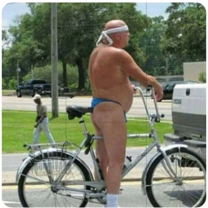 A shirtless, bald man wearing blue underwear and a white headband stands on a bicycle, facing away from the camera. The…