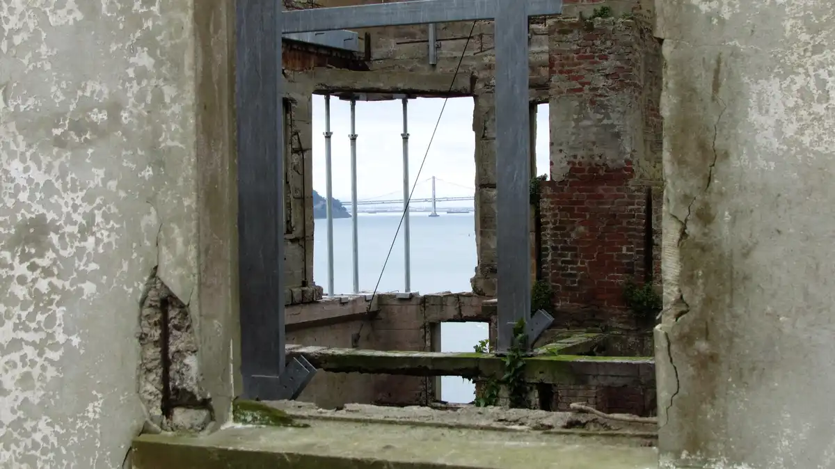 A view from inside a dilapidated, crumbling building looking out through a broken window frame at a distant suspension…