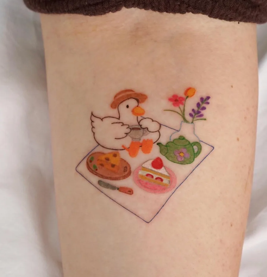 A tattoo on a person's leg depicts a cartoon duck wearing a hat, sitting at a picnic blanket with a teapot, a slice of cake…