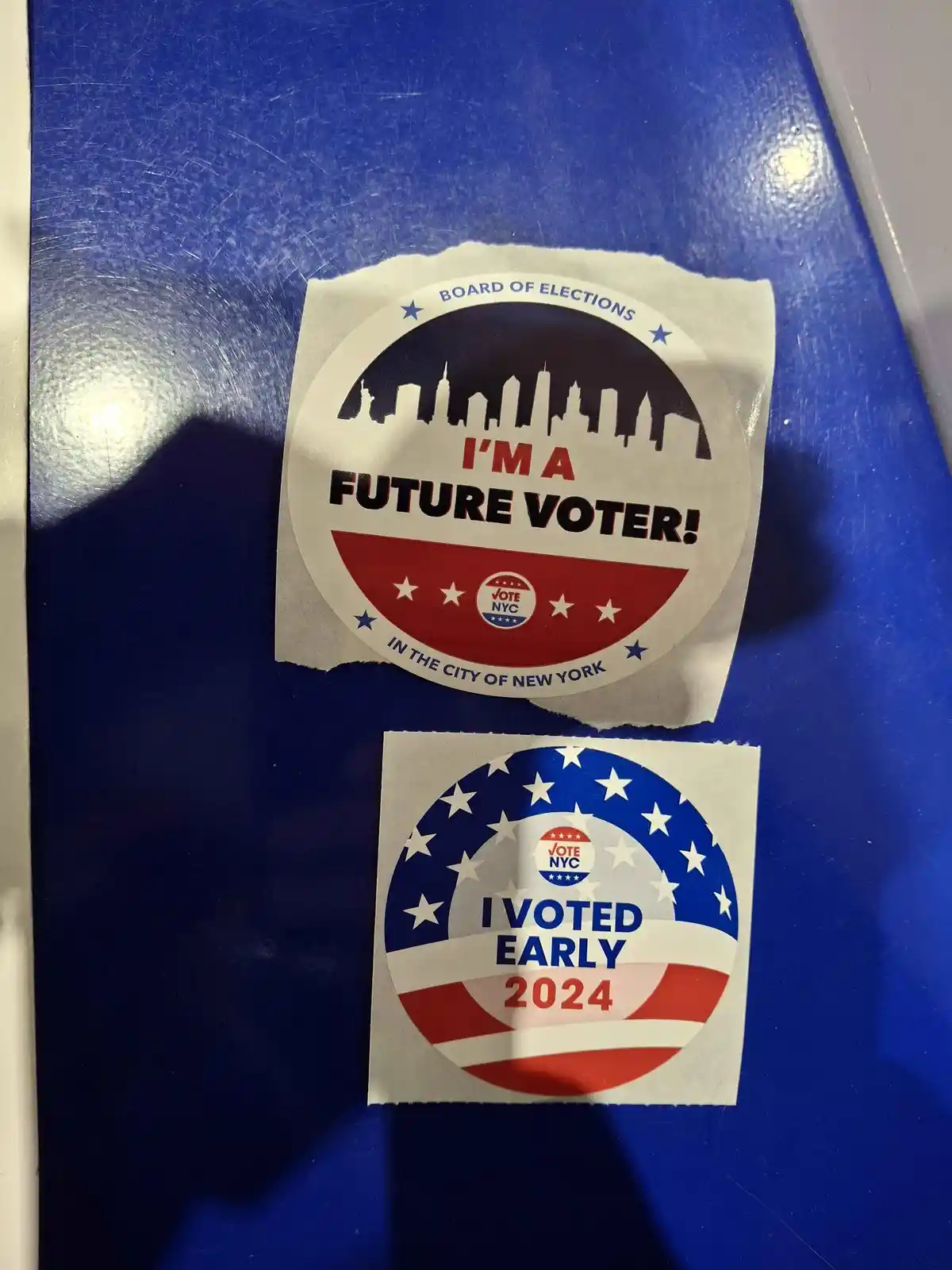 Two official-looking New York City election stickers are stuck on a blue surface. The top sticker says 'I'M A FUTURE VOTER!'…