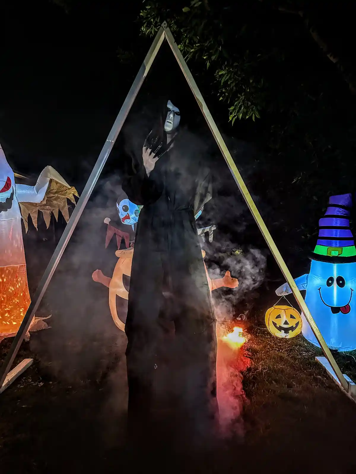 A person in a dark, smoky, Halloween-themed outdoor setup stands inside a large triangular frame, surrounded by inflatable…