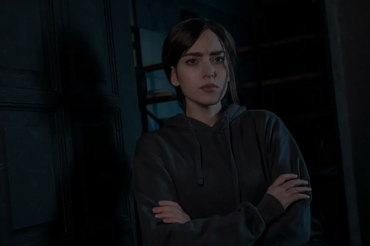 A woman with short dark hair wearing a dark hoodie stands in a dimly lit room with arms crossed, looking intensely at the…