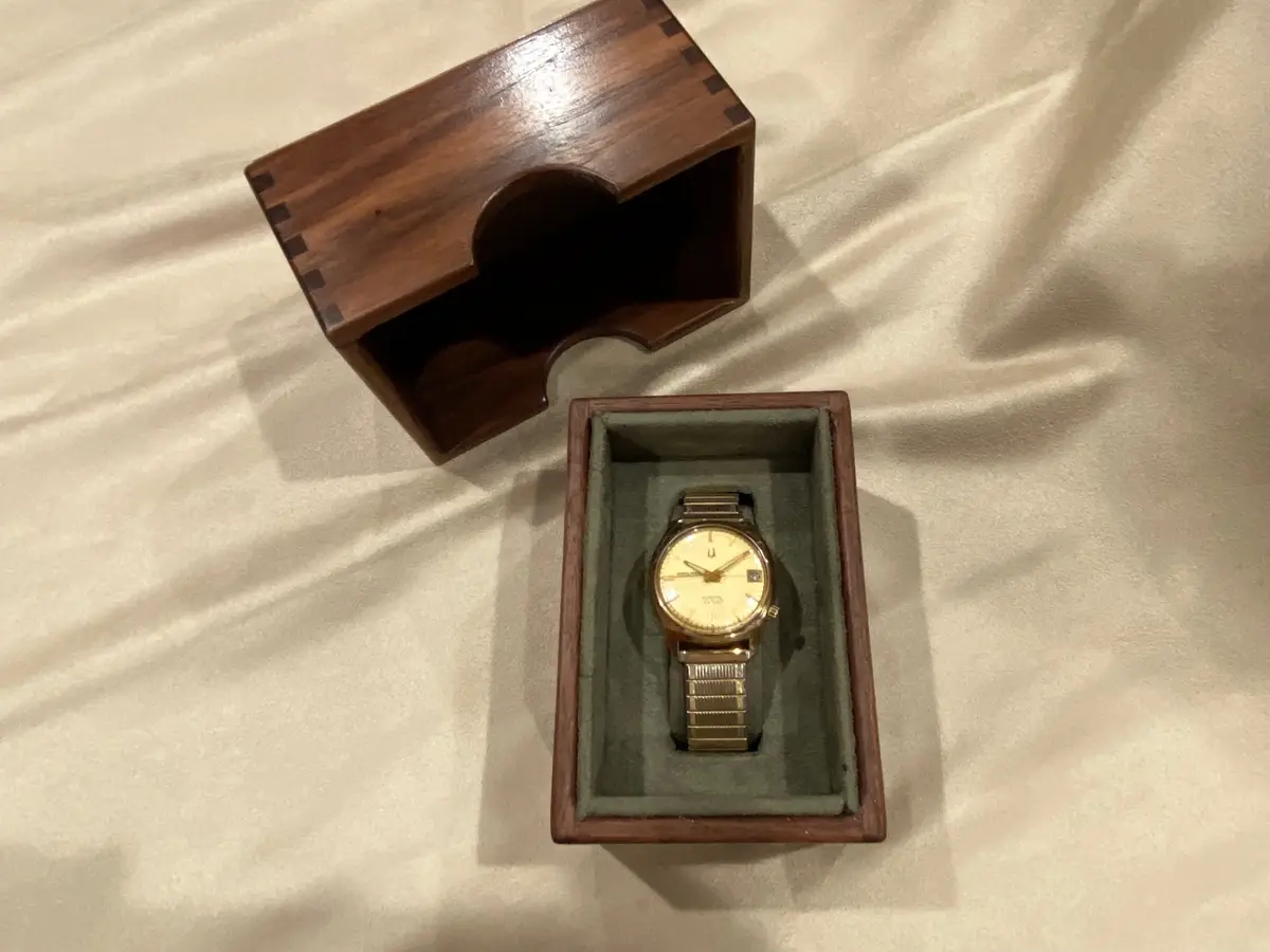 A gold-toned wristwatch with a metal bracelet sits in a dark green velvet-lined wooden presentation box, which is open on a…
