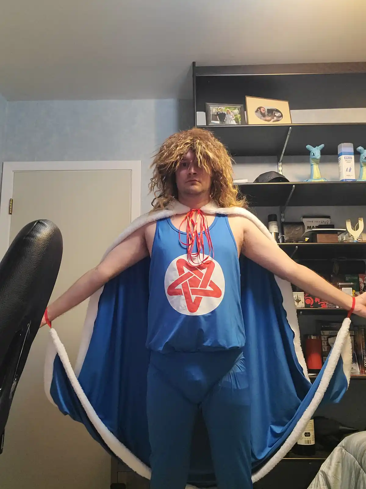 A person with a shaggy blonde wig poses indoors in a blue superhero costume with a white-trimmed cape, arms spread wide. The…