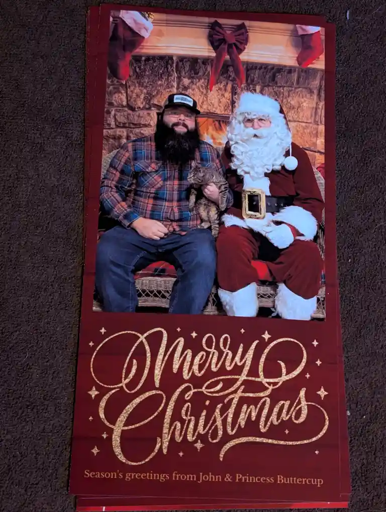 A Christmas card photo shows a man with a beard and plaid shirt holding a cat, sitting next to a person in a full Santa…