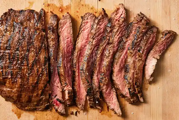 A top-down photo of a grilled steak sliced into thin pieces on a wooden cutting board, showing charred exterior and pink…
