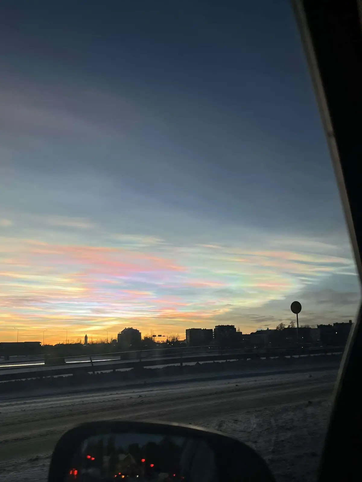 A photo taken from inside a car shows a stunning sunset with iridescent, rainbow-colored clouds, known as nacreous clouds…