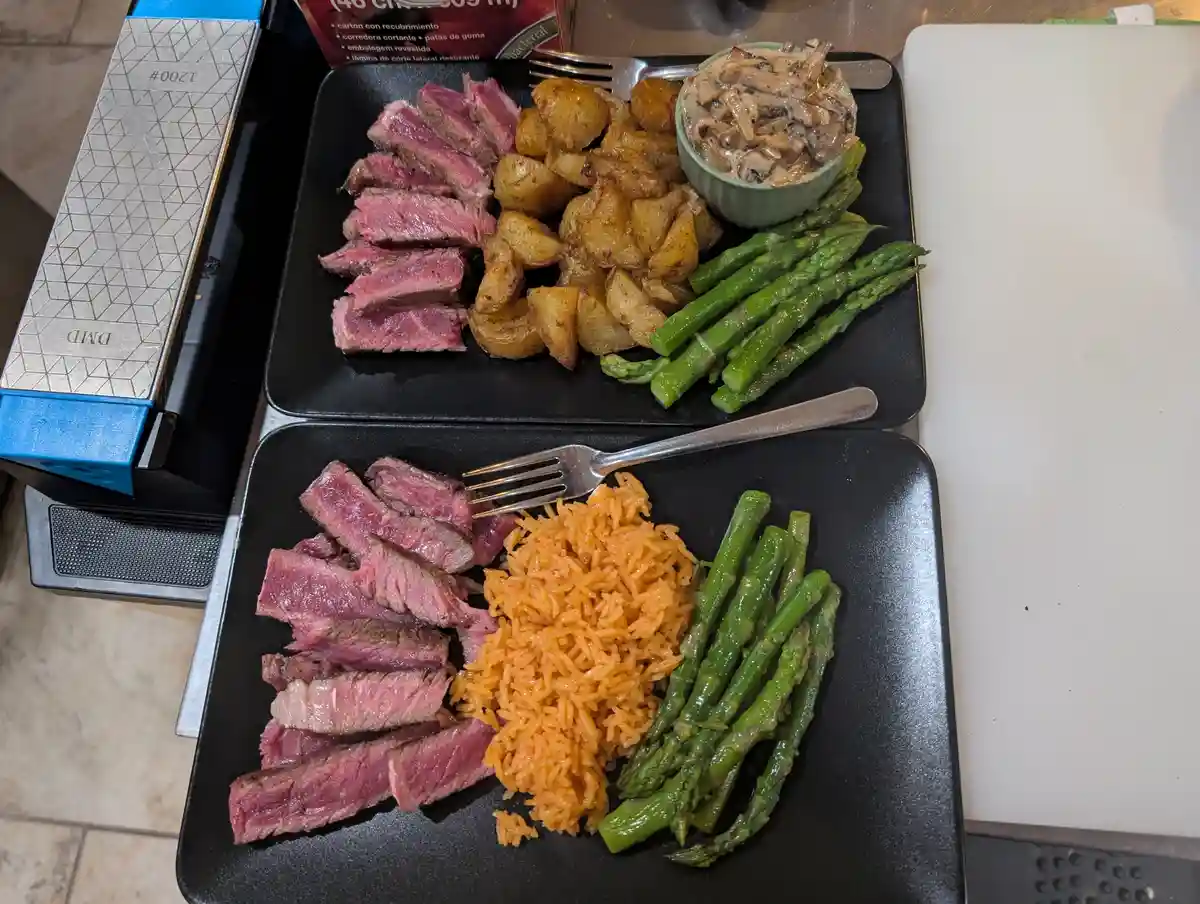 Two black plates show identical sliced steak and asparagus, but the top plate has roasted potatoes and mushroom sauce while…