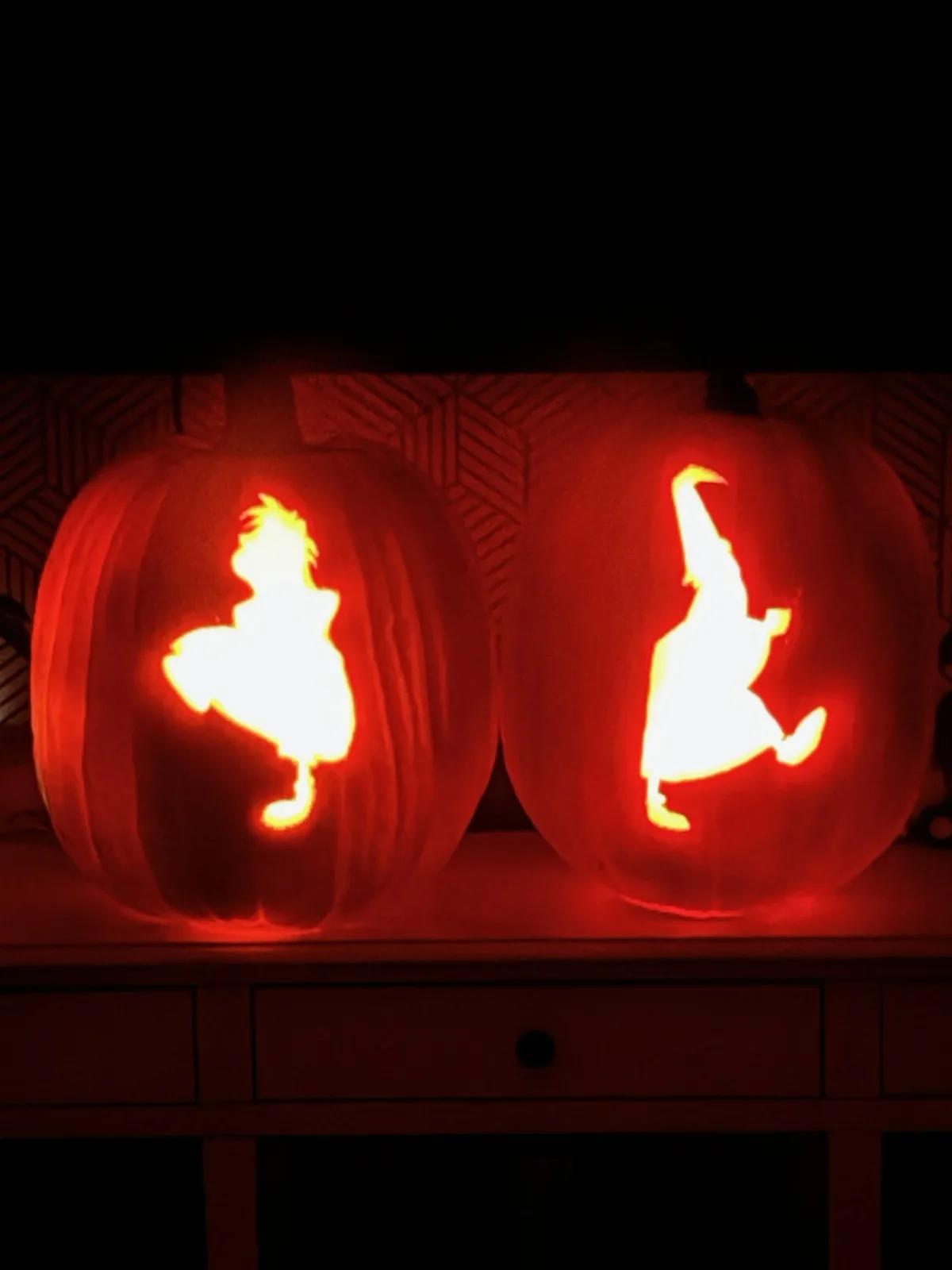 Two carved Halloween pumpkins sit side by side on a shelf, glowing from within. The left pumpkin features a silhouette of a…