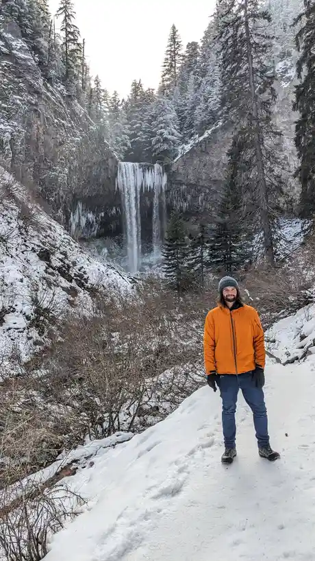 A man in an orange jacket, blue jeans, gloves, and a beanie stands smiling on a snowy trail in front of a partially frozen…